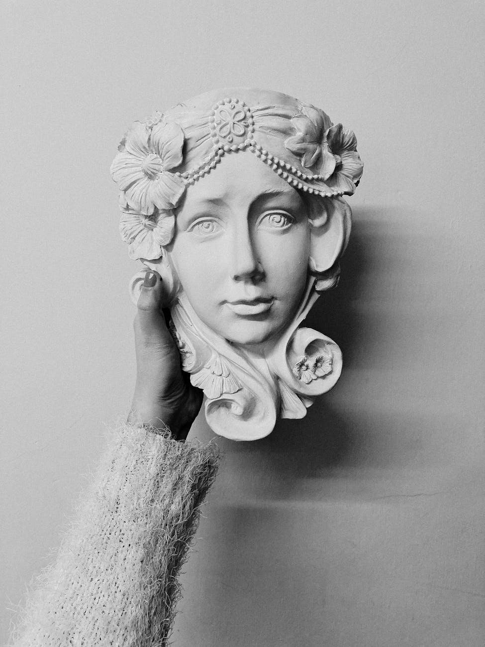 A grayscale photo of a black womans hand in a sweater holding aloft a plaster bust of a serene woman's face.