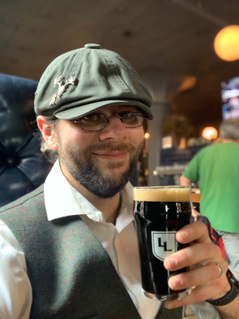 A selfie of the author wearing an olive green newsboy cap, green tweed vest, and white shirt raising a pint of dark beer.
