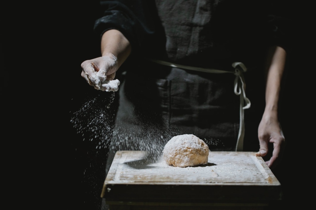 A baker in a black apron and with a black background dusts flour over a lump of dough on a small baking bench.